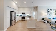This single-story residence in Irvine, California offers a thoughtfully designed open-concept layout with modern finishes throughout. The kitchen serves as a central hub, featuring white shaker cabinetry, stainless steel appliances, a gas range, and a large island with a contrasting wood-tone countertop, illuminated by pendant lighting and recessed fixtures. Light wood-style flooring flows seamlessly through the kitchen and living areas, enhancing the bright, airy atmosphere. The living room is well-proportioned, with a ceiling fan, sliding glass doors opening to an outdoor area, and ample natural light. A dedicated in-unit laundry closet with a stacked washer and dryer adds everyday convenience. The primary bedroom features soft blue accent walls, a natural wood headboard, wall-mounted sconce lighting, and large windows with garden views. The second bedroom offers neutral tones, plush carpet, a ceiling fan, and generous closet space. The primary bathroom includes a glass-enclosed walk-in shower with white subway tile and a teak stool, complemented by open built-in shelving. The second bathroom provides a full soaking tub with a tiled surround and additional open shelving for storage. Clean lines, a neutral palette, and contemporary finishes define this well-appointed Irvine home.
