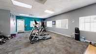 Located at 7650 Highlands View Rd in Truckee, California, this fitness center offers a thoughtfully appointed exercise room designed to support a well-rounded workout routine. The space is finished with durable carpet tile flooring and a drop ceiling with recessed panel lighting, creating a bright and functional environment. A bold teal accent wall adds a vibrant, energizing touch against the surrounding gray walls, giving the room a modern and inviting aesthetic. Natural light filters in through multiple windows fitted with white plantation shutters, contributing to the open, airy feel of the space.

The exercise room is comprehensively equipped with a variety of cardio machines, including a treadmill, elliptical trainer, and stationary spin bike, as well as a multi-station cable strength training machine by Hoist. Additional fitness accessories include stability balls, a punching bag, dumbbells, resistance tools, and a flat-screen TV mounted on the wall for entertainment during workouts. A dedicated stretch and floor exercise area rounds out the fitness options.

A built-in kitchenette along one wall features wooden cabinetry, countertop space, and small appliances including a microwave, providing convenient access to refreshments. A storage cabinet and organized shelving for free weights further enhance the room's practicality, making this facility a comprehensive and well-organized fitness destination.