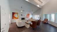 Located at 1237 Enclave Cir in Arlington, TX, this single-floor clubhouse offers an expansive and thoughtfully designed gathering space for residents. The centerpiece of the main lounge is a floor-to-ceiling tiled fireplace framed by vibrant, large-scale abstract artwork that adds bold pops of orange, red, and teal throughout the room. Soaring vaulted ceilings with exposed beams and pendant lighting create an airy, open atmosphere, while dark hardwood flooring grounds the space with warmth and contrast. The seating area is generously appointed with a curved cream sectional, classic leather wingback chairs, and a tufted ottoman-style coffee table, offering a variety of comfortable arrangements for socializing or relaxing. A row of large windows and glass doors along one wall floods the interior with natural light and provides direct sightlines to the outdoor pool area, seamlessly connecting the indoor and outdoor environments. Adjacent to the main lounge, a smaller dining nook features a round table with white chairs, ideal for casual gatherings or meetings. Multiple doorways lead to additional private rooms, adding functional versatility to the space. The overall ambiance is modern yet inviting, with a carefully curated design aesthetic that balances contemporary furnishings with vibrant artistic accents.
