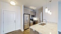 This single-floor residence at Overture showcases a thoughtfully designed open-concept layout that seamlessly connects the kitchen and living areas. The kitchen serves as a central highlight, anchored by a spacious island with light-toned quartz countertops featuring subtle veining. A double stainless steel sink with a contemporary arc faucet is integrated into the island, accompanied by a stainless steel dishwasher below. Light wood-grain cabinetry extends from floor to ceiling, providing generous storage, while stainless steel appliances — including a French door refrigerator, electric range, and over-the-range microwave — complete the cooking space. A mosaic tile backsplash adds texture behind the range. Three pendant lights with cylindrical glass shades hang above the island, enhancing both task lighting and visual appeal. The open living area beyond features wood-style flooring, neutral light gray walls, and a ceiling fan, creating a comfortable and airy atmosphere. A window allows natural light to fill the living space. The overall design aesthetic is clean, modern, and cohesive throughout.