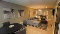 This single-floor space is designed with an open-concept layout that seamlessly connects its key functional areas. The living area serves as the centerpiece, anchored by a large gray sectional sofa with a matching ottoman, all resting atop a patterned area rug over light hardwood flooring. A dark leather accent chair and a round side table add sophistication to the seating arrangement. Abstract artwork adorns the warm taupe-toned walls, contributing to the space's contemporary aesthetic.

Adjacent to the living area, a compact dining setup features a round dark table paired with sleek gray chairs, providing an intimate space for meals or gatherings. Recessed lighting throughout the ceiling ensures consistent, warm illumination across the open floor plan.

Toward the rear of the space, a kitchenette or wet bar area is visible, complete with dark cabinetry, a countertop, and a sink. An oval mirror above the counter adds a decorative touch. A doorway leads to an additional room, hinting at further functional space beyond the main living area. Overall, the design balances modern style with comfort through cohesive furnishings and a neutral color palette.