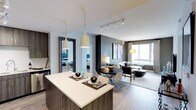 This thoughtfully designed single-floor residence showcases a seamless open-concept layout that connects the kitchen, dining, and living areas into one cohesive and inviting space. The kitchen features a large island with a crisp white quartz countertop and rich wood cabinetry, illuminated by a pair of elegant pendant lights that serve as a stylish focal point. Track lighting overhead further enhances the modern aesthetic while ensuring the space is well-lit throughout. The dining area accommodates a round table with contemporary seating, positioned naturally between the kitchen and living room to encourage effortless flow. The living room is bright and airy, benefiting from expansive windows that draw in abundant natural light and provide views of the surrounding area. A neutral color palette of whites, grays, and warm wood tones creates a clean, sophisticated ambiance throughout. Light hardwood-style flooring extends across the main living areas, adding warmth and continuity. A hallway visible from the main space leads to additional rooms, including a bedroom appointed with its own refined finishes. The overall design balances form and function, offering a contemporary and comfortable environment with carefully curated furnishings and decor.