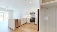 This thoughtfully designed one-bedroom, one-bathroom residence offers an efficient and functional single-floor layout with modern finishes throughout. The kitchen is a standout feature, showcasing crisp white shaker-style cabinetry extending to the ceiling, a classic subway tile backsplash, light quartz countertops, and a full suite of stainless steel appliances including a dishwasher, range, microwave, and refrigerator. A large kitchen island with a built-in sink and bar-height legs provides additional prep space and casual seating, seamlessly connecting the kitchen to the open living area. Light-toned hardwood flooring flows throughout the main living spaces, contributing to a bright and airy ambiance complemented by recessed lighting and tall white walls. The bedroom is a calm, carpeted retreat featuring neutral tones, a window with views to the outdoors, and ample room for furnishings. The bathroom maintains the home's clean aesthetic with white cabinetry, a quartz vanity top, wood-tone flooring, and a glass-enclosed walk-in shower with marble-style tile surround. Overall, this residence presents a cohesive, contemporary design with a light-filled atmosphere and practical, well-appointed spaces.