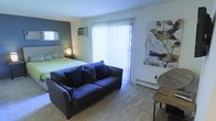 This single-floor studio efficiently combines a sleeping and living area into one open layout. The space features a large sliding glass door dressed with vertical blinds, allowing abundant natural light to fill the room and providing potential access to an outdoor area. The living zone centers around a navy blue sofa accented with patterned throw pillows, positioned on a carpeted floor that adds warmth underfoot. A small dining or side table is tucked to one corner, adding functional utility. The sleeping area is defined by a blue-gray accent wall, which adds depth and visual distinction to the room. A wall-mounted air conditioning unit serves the space, complemented by a baseboard heater near the sliding door for year-round climate control. Abstract wall art adds a decorative focal point, enhancing the room's character. A bedside lamp provides ambient lighting for the sleeping zone. The overall ambiance is casual and practical, with a cool, relaxed color palette of blues and neutrals creating a cohesive feel throughout the combined living and sleeping space.