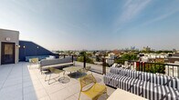Located in Philadelphia's vibrant Fishtown neighborhood, this rooftop terrace at 1901 E York St is a generously sized outdoor amenity space that delivers an elevated urban experience. The terrace is divided into two distinct zones connected by a hallway, together offering a well-rounded variety of seating and gathering areas. Large-format terrazzo-style tile flooring runs throughout, lending a clean, contemporary aesthetic underfoot.

The first zone features a lounge area furnished with bold striped sectional sofas, wire-frame accent chairs in gold, and low coffee tables — ideal for relaxed social gatherings. This section is partially sheltered by the building's corrugated metal façade, which provides a backdrop of modern industrial character. A striking wood-and-steel pergola anchors a second area, providing partial shade and architectural interest while framing views of the surrounding rooftops.

The second zone is configured for dining and casual seating, with bistro-style tables, red accent chairs, and bar-height tables with dark seating. Black metal railings line the perimeter throughout, ensuring safety while preserving unobstructed panoramic views of the Philadelphia skyline — most notably the Center City high-rises visible in the distance. Large sliding glass doors connect the outdoor terrace to interior access points, completing this thoughtfully designed rooftop retreat.