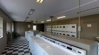 This single-floor laundry facility is designed to accommodate high-volume use with a practical and organized layout. A long row of top-load washing machines lines one side of the room, offering ample capacity for simultaneous loads. Opposite, a full wall of stacked dryer units provides an equally generous number of drying options, maximizing the room's efficiency. The classic black-and-white checkered floor adds a timeless visual contrast to the clean, white appliances throughout the space. Overhead, fluorescent lighting runs along the ceiling in evenly spaced fixtures, ensuring the room is brightly and uniformly lit. The walls feature warm wood-toned paneling, lending a subtle sense of warmth to the otherwise utilitarian setting. Support columns are integrated neatly into the layout without disrupting the flow of the space. A countertop surface provides a convenient area for sorting and folding laundry. Informational postings are visible near the entrance, keeping users informed of facility guidelines. Overall, the space is clean, functional, and well-maintained, reflecting a straightforward design focused on practicality and ease of use.