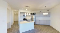 This contemporary single-floor residence is defined by its clean, modern aesthetic and thoughtfully designed layout. The heart of the home is an open-concept kitchen that seamlessly integrates style and functionality. A central island with a light quartz countertop and an undermount sink serves as both a prep area and a casual gathering point, illuminated by elegant crystal pendant lights overhead. The kitchen is outfitted with a full suite of stainless steel appliances, including a gas range with microwave above and a French door refrigerator, all complemented by sleek grey flat-panel cabinetry. A distinctive built-in wine rack adds a refined touch to the cabinetry wall. Recessed lighting throughout ensures the space is bright and well-lit, while a secondary pendant fixture highlights an additional countertop zone, ideal for dining or entertaining. Wide-plank wood-style flooring runs throughout, lending warmth and continuity to the open layout. The space flows naturally toward adjacent living areas, with a hallway leading to further rooms beyond. The overall ambiance is light, airy, and modern, offering a welcoming environment with a polished, move-in-ready feel.