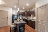 Located in Sandy, Utah, this single-floor residence offers a thoughtfully designed open-concept layout that seamlessly connects the kitchen and living area. The kitchen is a standout feature, equipped with rich dark-espresso cabinetry, granite countertops, a central island with bar seating, pendant lighting, and black appliances including a full-size refrigerator, range, microwave, and dishwasher. Hardwood-style flooring runs throughout the main living area, adding warmth and continuity to the space. The living area is generously sized, bathed in natural light from multiple windows, and offers a comfortable, open atmosphere well-suited for both relaxing and entertaining. The bedroom is a calm retreat, finished with plush carpeting, a ceiling fan, natural light from a large window, and a warm neutral accent wall. An adjacent full bathroom features tile flooring, a granite vanity, and a tub-shower combination. The overall design palette of warm neutrals, accented by the dark cabinetry and hardwood tones, creates a cohesive and inviting ambiance throughout the home.