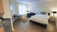 This single-floor space centers around a generously sized bedroom that combines comfort and functionality in a clean, modern layout. The room features warm hardwood flooring throughout, which contrasts nicely with the light-toned walls to create a bright, welcoming atmosphere. A large window fitted with white horizontal blinds allows natural light to filter in, illuminating the space evenly.

The sleeping area is anchored by a king-sized bed dressed in neutral white bedding accented with teal and gray decorative pillows. Adjacent to the bed, a cozy seating area includes a deep navy blue loveseat and a coordinating accent chair, arranged around a colorful floral area rug. A cheerful yellow ottoman serves as a central accent piece, adding a pop of color to the arrangement.

A sleek glass-topped desk with a slim metal frame is positioned near the window, accompanied by a small potted plant, providing a practical workspace within the room. A floor lamp adds warm ambient lighting to the seating area. The overall ambiance is comfortable and stylish, blending practical furnishings with thoughtful decorative touches.