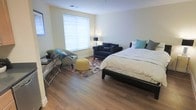 This single-floor space centers around a generously sized bedroom that combines comfort and functionality in a clean, modern layout. The room features warm hardwood flooring throughout, which contrasts nicely with the light-toned walls to create a bright, welcoming atmosphere. A large window fitted with white horizontal blinds allows natural light to filter in, illuminating the space evenly.

The sleeping area is anchored by a king-sized bed dressed in neutral white bedding accented with teal and gray decorative pillows. Adjacent to the bed, a cozy seating area includes a deep navy blue loveseat and a coordinating accent chair, arranged around a colorful floral area rug. A cheerful yellow ottoman serves as a central accent piece, adding a pop of color to the arrangement.

A sleek glass-topped desk with a slim metal frame is positioned near the window, accompanied by a small potted plant, providing a practical workspace within the room. A floor lamp adds warm ambient lighting to the seating area. The overall ambiance is comfortable and stylish, blending practical furnishings with thoughtful decorative touches.
