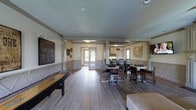 This inviting game room is designed to accommodate a variety of social and recreational activities, all within a single, open-concept floor. The space is finished with warm hardwood-style flooring and neutral-toned walls accented by classic wainscoting, giving the room a refined yet casual atmosphere. Recessed lighting throughout ensures the space is well-lit and welcoming.

A standout feature is the full-size shuffleboard table positioned along one wall, accompanied by decorative wall art that adds a lively, entertainment-focused character to the room. Multiple high-top dining tables with upholstered chairs are arranged throughout the space, providing ample seating for gatherings and social events.

Structural columns define distinct zones within the open layout without compromising the sense of openness. Toward the rear of the space, a kitchenette with white cabinetry offers functional support for hosting, making it easy to prepare and serve refreshments. A wall-mounted television is also present, adding to the entertainment options available.

Natural light filters in through a glass-paneled door at the back, connecting the interior to an exterior entry point. Decorative wall hangings and artwork complete the space, lending personality and warmth. Overall, this game room is a versatile, thoughtfully designed communal space ideal for recreation and social gatherings.