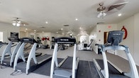 This fitness center offers a comprehensive workout environment designed to meet a variety of exercise needs. The space is outfitted with multiple treadmills from True Fitness, elliptical machines, and an assortment of strength training equipment, providing users with a full range of cardio and resistance training options. The layout is open and well-organized, allowing for comfortable movement between machines. Recessed lighting throughout the ceiling ensures the space is well-lit, while ceiling fans contribute to air circulation and comfort during workouts. The textured ceiling and clean white walls give the room a classic, tidy appearance. Toward the far end of the space, an arched window allows natural light to filter in, adding warmth and an inviting ambiance to the environment. Motivational wall decals are visible, contributing to an encouraging atmosphere. The flooring features durable carpet suitable for a fitness setting. A doorway at the rear provides additional access or connectivity to adjacent areas. Overall, the fitness center is practical, well-maintained, and thoughtfully arranged to support an active and healthy lifestyle.