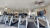 This fitness center offers a comprehensive workout environment designed to meet a variety of exercise needs. The space is outfitted with multiple treadmills from True Fitness, elliptical machines, and an assortment of strength training equipment, providing users with a full range of cardio and resistance training options. The layout is open and well-organized, allowing for comfortable movement between machines. Recessed lighting throughout the ceiling ensures the space is well-lit, while ceiling fans contribute to air circulation and comfort during workouts. The textured ceiling and clean white walls give the room a classic, tidy appearance. Toward the far end of the space, an arched window allows natural light to filter in, adding warmth and an inviting ambiance to the environment. Motivational wall decals are visible, contributing to an encouraging atmosphere. The flooring features durable carpet suitable for a fitness setting. A doorway at the rear provides additional access or connectivity to adjacent areas. Overall, the fitness center is practical, well-maintained, and thoughtfully arranged to support an active and healthy lifestyle.