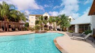 This property showcases a well-appointed outdoor amenity area centered around a generously sized, curved swimming pool with striking turquoise water. A decorative fountain serves as a focal point within the pool area, adding an elegant, resort-like atmosphere to the space. The pool deck is finished with stone-toned tiling and features ample space for lounging, with outdoor seating conveniently positioned nearby. Tall, mature palm trees are thoughtfully planted throughout the grounds, lending a tropical ambiance and providing natural shade across the outdoor space. The surrounding landscaping is neatly maintained, with manicured shrubs and ornamental plantings enhancing the overall aesthetic. In the background, the multi-story building presents a warm stucco facade with neutral tan tones, gabled rooflines, and multiple windows and balconies that overlook the pool area. A covered pergola structure is visible to one side, offering additional shaded outdoor space. The combination of the lush landscaping, sparkling pool, and inviting architectural design creates a cohesive and attractive outdoor living environment with a distinctly warm-climate, leisure-oriented character.