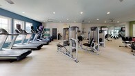 Located at 1500 Parklawn Dr in Charleston, South Carolina, this single-level fitness center is designed to support a comprehensive workout experience. The space features light hardwood-style flooring throughout, recessed lighting, and ceiling fans that contribute to a bright and comfortable environment. Accent walls in sage green and navy blue add a modern, energizing aesthetic to the interior.

The facility is divided into distinct workout zones. A dedicated cycling and stretching area is anchored by large floor-to-ceiling mirrors on both sides, with natural light streaming in through shuttered windows overlooking the surrounding landscape. Stability balls and resistance equipment are neatly stored in this section for flexibility and core training.

The main floor houses an extensive array of strength training machines, including cable and weight resistance equipment, as well as a dumbbell rack with a full range of free weights. A row of treadmills and elliptical machines lines one side of the room, each equipped with individual screens for entertainment during cardio sessions. Mounted flat-screen TVs are positioned throughout for added convenience.

Ample open floor space between equipment zones allows for ease of movement and additional floor exercises. The overall layout is thoughtfully organized, making the fitness center accessible and functional for users of all fitness levels.