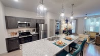 Located at 11701 Mira Lago Blvd in Farmers Branch, Texas, this thoughtfully designed single-floor residence offers a seamless blend of modern finishes and functional living spaces. The kitchen serves as a centerpiece of the home, showcasing dark charcoal cabinetry, granite countertops, a generously sized island with an undermount sink and bar seating, and a full suite of stainless steel appliances. Stylish pendant lighting overhead adds a refined touch to the space. Light wood-style flooring flows throughout the main living areas, creating a cohesive and contemporary aesthetic. The entryway provides convenient access to a pantry closet and opens naturally into the kitchen and adjacent living areas. The living room is bright and inviting, featuring a ceiling fan, natural light from a well-placed window with sheer drapery, and ample space for comfortable seating arrangements. The bedroom is a restful retreat, complete with carpet flooring, a ceiling fan, and windows dressed with layered striped curtains that allow for both privacy and natural light. Each bathroom is finished with granite-topped dark vanities and wood-style flooring, with one bathroom offering direct access to a spacious walk-in closet. Throughout the residence, neutral tones and clean architectural lines create a polished, move-in-ready environment.