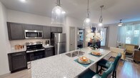 Located at 11701 Mira Lago Blvd in Farmers Branch, Texas, this thoughtfully designed single-floor residence offers a seamless blend of modern finishes and functional living spaces. The kitchen serves as a centerpiece of the home, showcasing dark charcoal cabinetry, granite countertops, a generously sized island with an undermount sink and bar seating, and a full suite of stainless steel appliances. Stylish pendant lighting overhead adds a refined touch to the space. Light wood-style flooring flows throughout the main living areas, creating a cohesive and contemporary aesthetic. The entryway provides convenient access to a pantry closet and opens naturally into the kitchen and adjacent living areas. The living room is bright and inviting, featuring a ceiling fan, natural light from a well-placed window with sheer drapery, and ample space for comfortable seating arrangements. The bedroom is a restful retreat, complete with carpet flooring, a ceiling fan, and windows dressed with layered striped curtains that allow for both privacy and natural light. Each bathroom is finished with granite-topped dark vanities and wood-style flooring, with one bathroom offering direct access to a spacious walk-in closet. Throughout the residence, neutral tones and clean architectural lines create a polished, move-in-ready environment.