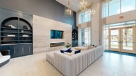 Located at 14000 Tapestry Dr in Orlando, FL, this sophisticated single-floor space offers a generously sized open layout designed with a keen attention to architectural detail and contemporary style. The living area serves as the centerpiece of the space, anchored by a striking light wood accent wall that houses a sleek linear fireplace and a wall-mounted television, flanked by dark navy built-in shelving units with arched detailing. Soaring double-height ceilings amplify the sense of openness, while dramatic gold geometric chandeliers add a refined, upscale ambiance. Large floor-to-ceiling windows dressed in sheer white drapery flood the interior with abundant natural light, offering views to the outdoors. Light-toned large-format tile flooring runs throughout, lending a clean and cohesive aesthetic. A dedicated lounge area features a sculptural vertical wood slat partition that serves as an architectural focal point, complemented by marble-top round tables and curved upholstered seating. A separate casual gathering area includes a bar-height counter with dark cabinetry, pendant lighting, and additional seating, ideal for socializing. Curated artwork and woven accent chairs add warmth and personality throughout the space.