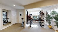 Located at 11126 SE 256th St in Kent, Washington, this single-floor fitness center offers a thoughtfully designed workout facility suited for a comprehensive exercise routine. The space is divided into open, interconnected areas that flow seamlessly from one zone to the next, creating an inviting and functional environment.

The main workout area features durable, carpet-style flooring and is outfitted with a variety of cardio equipment, including a treadmill, elliptical trainer, and stationary spin bike. A multi-function strength training machine rounds out the equipment selection, providing options for a full-body workout. Large wall-mounted mirrors are strategically placed throughout, enhancing the sense of space and supporting proper form during exercise.

Natural light floods the interior through multiple large windows adorned with blinds, giving the space a bright and airy feel. Recessed ceiling lighting supplements the natural illumination throughout all areas. Decorative touches such as potted tropical plants, framed motivational artwork, and a water cooler add warmth and character to the otherwise functional setting.

A dedicated entryway area connects the zones and provides access to additional rooms, including what appears to be a restroom. A community bulletin board is also present, suggesting this facility serves a shared residential or community setting. Overall, the space strikes a balance between practical fitness functionality and a welcoming, well-maintained atmosphere.