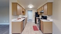 This single-floor unit offers a clean, functional layout with a cohesive neutral color palette and durable finishes throughout. The open living area features light-toned luxury vinyl plank flooring, white trim, and multiple windows fitted with blinds that allow natural light to fill the space. A full-light exterior door provides additional brightness and outdoor views of surrounding greenery. A baseboard heater runs along one wall, ensuring efficient climate control.

The galley-style kitchen is efficiently designed with warm wood-toned cabinetry lining both sides, offering generous storage and counter space. White countertops complement the stainless steel sink, dishwasher, and refrigerator, while a black electric range with an overhead exhaust hood completes the appliance suite. The kitchen flooring continues the vinyl plank theme, and a doorway at the far end connects to the rest of the unit.

The bedrooms feature soft carpeting in neutral tones, providing a comfortable underfoot feel. Each room includes windows with blinds and clean, understated wall colors in warm beige and gray tones. A separate room with darker vinyl plank flooring offers flexible use as a living space or office.

The full bathroom is equipped with a single-sink vanity featuring wood cabinetry, a large mirror, and a tub-shower combination. Light vinyl plank flooring ties the bathroom together with the rest of the unit's consistent design aesthetic.