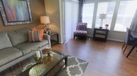 This well-appointed single-floor residence offers a seamlessly designed living area that balances comfort and contemporary style. The space is anchored by rich hardwood flooring that flows throughout, lending warmth and visual continuity to the interior. Large windows dressed with elegant drapes and blinds flood the room with natural light, creating a bright and airy atmosphere.

The living area is tastefully furnished with a plush grey sofa accented by vibrant orange and patterned throw pillows, adding a pop of color to the neutral palette. A sleek glass-top coffee table serves as a functional centerpiece, complemented by a patterned area rug in soft grey tones. A chic accent chair upholstered in a bold purple geometric print adds personality and visual interest to the space.

Thoughtful lighting is provided by stylish table lamps positioned throughout the room, enhancing the warm, inviting ambiance during evening hours. The neutral wall tones provide a versatile backdrop that ties together the eclectic mix of furnishings and décor. Vertical blinds at the sliding door offer both privacy and light control while maintaining the room's clean, modern aesthetic. Overall, the space exudes a curated, welcoming charm suited for both relaxing and entertaining.