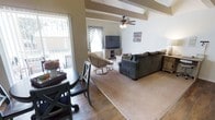 This single-story home showcases a thoughtfully designed open-concept layout that connects the dining and living areas into one cohesive, comfortable space. The dining area features rich hardwood-style flooring and is furnished with a classic dark wood table and matching chairs, positioned near a large window that invites an abundance of natural light. A sliding glass door provides direct access to an outdoor area, enhancing the sense of openness and connectivity to the exterior.

The living room flows naturally from the dining space, transitioning to plush carpeting that adds warmth underfoot. Vaulted ceilings with exposed decorative beams give the room a sense of generous vertical space, while a ceiling fan with an integrated light fixture provides both comfort and ambient illumination. The living area is anchored by a comfortable sectional sofa and complemented by rattan accent chairs, creating a relaxed, welcoming atmosphere. A media console with a flat-screen television serves as a functional focal point. The combination of neutral tones, natural textures, and ample light throughout gives the home a warm and inviting ambiance.