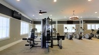 This fitness center offers a thoughtfully designed workout environment that balances functionality with a clean, modern aesthetic. The space is anchored by a large multi-station cable and strength training machine positioned at the center, providing a versatile range of resistance exercises for users of all fitness levels. Flanking the strength equipment is a dedicated cardio zone outfitted with treadmills and elliptical machines, arranged to maximize floor space and ease of movement between stations.

Natural light floods the room through multiple windows dressed with white blinds, creating an open and energizing atmosphere. The walls feature a two-tone paint scheme, with a warm neutral lower half contrasted by a deep charcoal upper band, lending the space a polished and contemporary feel. Crown molding adds a refined architectural touch.

Overhead, a ceiling fan with a sleek dark finish promotes air circulation throughout the room, complementing the recessed lighting that ensures consistent, even illumination. An elegant circular chandelier adds a distinctive decorative element, elevating the ambiance beyond a typical utilitarian gym setting. The light-toned carpeted flooring provides comfort underfoot while maintaining a cohesive interior design. Overall, this fitness center delivers a welcoming, well-appointed environment suited for a full-body workout routine.