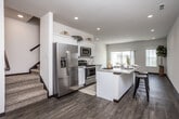 This two-story residence is designed with a clean, contemporary aesthetic that balances functionality and comfort throughout both levels. The main floor centers around an open-concept layout where the kitchen and living area flow seamlessly together. The kitchen is well-appointed with white shaker-style cabinetry extending to the ceiling, stainless steel appliances including a French door refrigerator, gas range, and built-in microwave, along with a central island featuring granite-style countertops and bar seating. Rich, wood-style plank flooring runs throughout the main level, complementing the recessed lighting and neutral wall tones. A sliding glass door in the living area opens to a private balcony, inviting natural light into the space. A half bathroom with a white vanity and large mirror is also found on this level. A carpeted staircase leads to the upper floor, where the bedrooms are finished with plush carpet, ceiling fans, and windows fitted with vertical blinds. The upper level also includes bathrooms outfitted with standard fixtures, including a tub-shower combination and vanity. Overall, the home presents a bright, neutral palette with modern finishes throughout.