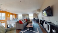 This dynamic, single-floor space makes a bold visual statement through its striking combination of vivid orange accent walls and neutral warm tones, creating an energetic yet balanced atmosphere throughout. Rich hardwood flooring runs continuously across the entire level, grounding the open layout with warmth and cohesion.

The main living area is generously proportioned, anchored by a large sectional sofa with colorful throw pillows, a wood-and-metal coffee table, and a bold geometric area rug. A wall-mounted flat-screen television sits atop a sleek dark media console, offering an organized entertainment focal point. Vaulted ceilings with angled planes add architectural interest and a sense of vertical volume to the room.

A dedicated workspace with a dark desk and office seating is seamlessly integrated into the open layout, bordered by large windows that flood the area with abundant natural light and offer views of the surrounding outdoor scenery. A dining area with a round table and green chairs occupies a well-lit corner adjacent to the boldly colored walls.

A prominent feature of the space is the white-railed mezzanine balustrade that lines the open interior overlook, connecting the various functional zones while maintaining visual openness. A ceiling fan provides additional airflow throughout. Multiple large windows on all sides ensure the space remains bright and connected to the outdoors throughout the day.