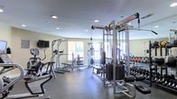 This fitness center offers a thorough and well-organized workout environment designed to accommodate a wide range of exercise routines. The space is outfitted with an impressive array of strength training equipment, including a multi-station cable machine, a power tower, a weight bench, and a full dumbbell rack stocked with a variety of free weights. Cardio options are also well represented, with machines such as an elliptical trainer visible alongside other aerobic equipment. A wall-mounted flat-screen television adds an entertainment element to complement workout sessions. The room benefits from large windows that invite natural light, creating a bright and energizing atmosphere. Recessed ceiling lights provide consistent, even illumination throughout the space. The flooring consists of durable, dark-toned rubber or sport-grade material suited for heavy equipment and high-impact activity. Full-length mirrors line a portion of the walls, enhancing the sense of spaciousness while allowing users to monitor their form. The walls are finished in a neutral, light tone that keeps the environment feeling clean and open. Overall, the fitness center is thoughtfully arranged to maximize usable floor space while maintaining clear pathways between equipment stations.