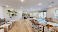 Located at 2920 Frontage Rd in Gainesville, Georgia, this single-floor living space offers a generously proportioned open layout finished with wide-plank hardwood flooring throughout. The room is bathed in natural light from large windows and expansive sliding glass doors that open onto a private balcony, providing serene views of the surrounding wooded landscape.

The interior design embraces a bohemian-inspired aesthetic, combining natural textures and organic elements with modern touches. A standout feature is the ceiling-suspended hammock chairs, which add a distinctive, relaxed character to the space. Layered area rugs define distinct lounging zones, complemented by cushioned seating, woven accent chairs, and low-profile coffee tables. A wall-mounted flat-screen television anchors one end of the room, while the opposite wall is adorned with rustic wooden shutter panels, wall-mounted planters overflowing with lush greenery, and gold-toned fixtures that add warmth to the crisp white walls.

A secondary seating nook features a low, cushioned sofa with a decorative rattan peacock headboard and a row of clean white accent tables — a cozy retreat within the larger room. Recessed ceiling lighting provides even illumination throughout, enhancing the airy, welcoming ambiance of this thoughtfully designed communal living space.