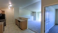 This single-floor space is designed with an open-concept layout that seamlessly connects the kitchen and living area. The kitchen features light wood cabinetry, a granite-style countertop, a stainless steel dishwasher, and a double basin sink. Tile flooring grounds the kitchen space, providing a practical and easy-to-maintain surface. A half-wall peninsula separates the kitchen from the adjoining living area, adding definition while maintaining an open feel. The living area transitions to plush wall-to-wall carpeting, creating a comfortable and inviting atmosphere. Recessed lighting in the kitchen provides focused illumination, while natural light filters in through a window visible in the living area, brightening the space. The neutral wall tones throughout contribute to a clean, versatile aesthetic. A baseboard heater along the living area wall ensures efficient climate control. Additional doors visible from the main living space suggest access to further rooms or storage, adding to the overall functionality of this well-organized, single-level layout.