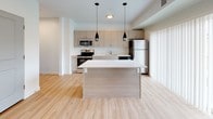 This single-floor residence is designed with a clean, contemporary aesthetic throughout. The open-concept kitchen serves as the centerpiece of the living area, featuring light wood-tone cabinetry, white quartz countertops, and a full suite of stainless steel appliances including a range, microwave, dishwasher, and refrigerator. A generously sized kitchen island with a wood-paneled base provides additional prep and seating space, illuminated by two sleek pendant lights overhead. Light hardwood flooring runs throughout the kitchen and common areas, lending a warm, cohesive feel.

The kitchen opens toward a sliding glass door framed by vertical blinds, offering access to an outdoor balcony and views of the surrounding landscape. This connection to natural light enhances the airy quality of the shared living space.

The bedroom is finished with plush, neutral-toned carpet and features a wide window that frames outdoor views, filling the room with natural light. The full bathroom, accessible via a bright hallway, is finished with hardwood-style flooring, a combination tub and shower, toilet, and a wood-toned vanity. The overall design of the unit prioritizes functional layout, natural light, and a neutral, modern finish palette throughout.