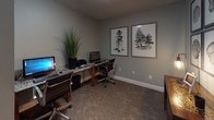 This business center is a thoughtfully designed, single-floor workspace that balances functionality with a polished aesthetic. The room is finished with cool gray walls and neutral-toned carpeting, creating a calm and focused environment well-suited for productivity.

The centerpiece of the space is a granite-topped workstation that accommodates multiple users, equipped with desktop computers, keyboards, and a printer, providing all essential tools for business tasks. Ergonomic rolling chairs with dark upholstery and chrome frames are positioned at each workstation, ensuring comfort during extended use.

Natural light filters into the room through a classic French door with divided-light panes, lending a welcoming brightness to the otherwise quietly lit interior. A wooden side table with a warm-toned mosaic surface and an industrial-style desk lamp adds a decorative touch near the entrance area.

The walls are adorned with framed nature-inspired artwork, including botanical tree illustrations and cross-section tree ring prints arranged in a grid pattern, contributing an organic, refined character to the space. Lush potted grass plants placed throughout add a fresh, natural element.

Clean white baseboards frame the room and complement the gray walls, while the overall layout remains open and uncluttered, offering a comfortable and professional environment for visitors needing a dedicated place to work.