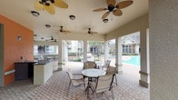 This inviting outdoor amenity space centers around a generously sized swimming pool framed by elegant brick paver decking, creating a polished and cohesive aesthetic throughout. The pool area is complemented by a spacious covered pavilion supported by sturdy stucco-finished columns, providing ample shade and comfort. Multiple tropical-style ceiling fans are mounted throughout the pavilion's ceiling, ensuring excellent airflow year-round. The pavilion also features an outdoor kitchen or bar counter, making it well-suited for gatherings and entertaining. Circular patio tables paired with sling-back chairs are arranged beneath the covered structure, offering comfortable seating options for residents or guests. Beyond the pavilion, the open pool deck is outfitted with sling lounge chairs and umbrella-shaded table sets, ideal for relaxing poolside. Mature trees and carefully maintained landscaping with layered shrubs and groundcover border the space, adding a lush, natural backdrop. The surrounding buildings display classic brick exteriors with large windows, pitched rooflines, and private balconies, lending a traditional architectural character to the community. The overall ambiance is that of a well-appointed, resort-inspired outdoor retreat designed for relaxation and social enjoyment.