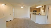 This single-floor residence offers a practical and comfortable layout centered around an open-concept living and kitchen area. The living space is generously sized, featuring neutral wall-to-wall carpeting that lends a warm, understated feel throughout. A ceiling fan with a light fixture provides both ventilation and ambient lighting, while a side window allows natural light to brighten the room. The kitchen is positioned in an open, semi-separated fashion, connected to the living area via a raised breakfast bar that doubles as a casual dining or prep surface. The kitchen is outfitted with warm wood-toned cabinetry offering substantial storage space, a gas range, and a built-in microwave above the stove. Under-cabinet lighting adds functional illumination to the workspace. The neutral color palette throughout — from the light beige walls to the matching carpet — creates a cohesive and move-in ready environment. The open flow between the kitchen and living area makes the space feel expansive and well-suited for both everyday living and entertaining. Overall, the residence presents a clean, functional interior with classic finishes and a welcoming atmosphere.