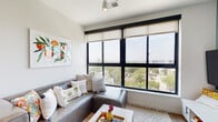 This contemporary single-floor residence is thoughtfully designed with a clean, modern aesthetic throughout. The living area is the standout centerpiece, featuring an expansive wall of floor-to-ceiling windows with bold black frames and roller shades, flooding the space with natural light and offering impressive elevated views of the surrounding cityscape and tree canopy. A grey sectional sofa, patterned area rug, and colorful wall art complete the inviting atmosphere.

The kitchen is well-equipped and stylish, showcasing white cabinetry with matte black hardware, light countertops, and a striking herringbone-patterned tile backsplash. Stainless steel appliances, including a range, microwave, and refrigerator, provide full functionality. A central island with seating offers additional prep space and casual dining, complemented by pendant lighting overhead and a vibrant teal accent wall.

The bedrooms are bright and comfortable, each featuring large windows with city views, light wood-toned flooring, and teal accent walls that add a cohesive, calming character. Each room includes space for a desk, making them functional for work or study.

The bathrooms are finished with wood-look flooring, wide vanity mirrors, white cabinetry, and soft blue-grey walls, creating a clean and serene environment. Throughout the unit, light hardwood flooring ties the spaces together with warmth and continuity.