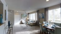 This single-floor space presents an open-concept layout that seamlessly connects the living and dining areas, creating a cohesive and functional environment. The living area is anchored by plush wall-to-wall carpeting in a neutral tone, complemented by soft grey walls that lend a calm, understated aesthetic throughout. A ceiling fan is centrally positioned overhead, adding both practicality and a clean architectural detail to the space.

Large windows fitted with grey draping panels allow generous natural light to filter into the room, brightening the interior and enhancing its sense of openness. The living area accommodates a comfortable sofa accented with blue and white throw pillows, paired with a patterned accent chair and a glass-topped coffee table, contributing to the light and airy feel. Decorative wall art in blue and neutral tones adds visual interest without overwhelming the space.

Adjacent to the living area, the dining space features a dark wood table surrounded by upholstered teal-toned chairs, providing an elegant contrast to the lighter surroundings. Recessed lighting supplements the natural light, ensuring the space remains well-lit at all hours. A white paneled entry door completes the layout, offering a clean and welcoming entryway into the home.