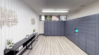 This single-level package room is designed to streamline parcel delivery and retrieval with a clean, functional layout. Dominating one wall is a large bank of Amazon Hub smart package lockers finished in slate gray. The lockers span nearly the full length of the room and feature an integrated touchscreen interface, allowing residents or users to retrieve deliveries securely at any time of day.

The flooring throughout the space consists of light wood-look vinyl planks, lending a warm, contemporary feel that complements the neutral white walls. Overhead, flush linear fluorescent fixtures provide consistent, bright illumination across the room. A row of clerestory windows along one wall allows natural light to filter in while maintaining privacy, offering glimpses of surrounding greenery.

One wall features vertical shiplap-style paneling painted in crisp white, adding a subtle textural element to the otherwise minimalist design. Decorative metallic wall sculptures are mounted symmetrically above a pair of sleek black console tables, which are accessorized with small potted plants and succulents. Framed botanical prints on an adjacent wall contribute to the tidy, polished ambiance. The overall atmosphere is modern, organized, and welcoming.