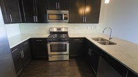This single-floor space showcases a thoughtfully designed interior with a strong emphasis on modern aesthetics and functional living. The kitchen stands out as a centerpiece, featuring rich dark cabinetry with sleek bar-pull hardware that creates a bold contrast against the light stone countertops. A full suite of stainless steel appliances — including a gas range with oven, an over-the-range microwave, and a double-basin sink with a contemporary arc faucet — equips the kitchen for everyday use and serious cooking alike. A subway tile backsplash in a soft, neutral tone adds texture and visual interest while maintaining a clean, polished look. The dark hardwood flooring runs throughout, grounding the space with warmth and continuity. The layout is efficient and well-organized, making excellent use of available space with ample upper and lower cabinet storage. The overall ambiance of the space is contemporary and cohesive, with a carefully curated palette of dark and light tones that gives the interior a sophisticated, urban feel. The attention to detail in the finishes and fixtures reflects a commitment to both style and practicality.