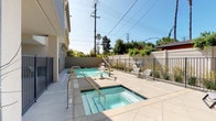 Located in the Westchester neighborhood of Los Angeles, this single-story outdoor amenity space at 5752 W 76th St offers a well-appointed pool and spa area designed for relaxation under the Southern California sun. The centerpiece is a generously sized rectangular swimming pool with clear, blue water, complemented by an adjacent hot tub spa, both finished with polished tile surrounds and equipped with stainless steel handrails for easy access.

The pool deck is surfaced in neutral-toned tile, providing a clean and modern aesthetic throughout. Comfortable lounge chairs are positioned poolside, ideal for sunbathing, while additional seating is available nearby. Large decorative planters with ornamental grasses and flowering plants add a refined, landscaped touch to the space.

The area is enclosed by a combination of iron fencing and perimeter walls, ensuring privacy and security, with a secured gate providing controlled access. Mature trees and lush greenery visible beyond the fencing contribute to a serene, garden-like atmosphere. The covered walkway adjacent to the pool features vaulted architectural detailing and tile flooring, connecting the outdoor amenities to the building's interior spaces. The overall ambiance is clean, modern, and well-maintained, reflecting a thoughtfully designed outdoor retreat.