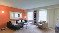 This single-floor space makes a strong first impression with its thoughtfully designed living room. A vibrant orange accent wall serves as a focal point, adding warmth and personality to the room, while a large framed mirror mounted on the wall enhances the sense of openness. The living area is generously sized, comfortably accommodating a charcoal sectional sofa, a sleek white coffee table, and a cushioned accent chair, all arranged to create an inviting and functional layout. Neutral carpet flooring runs throughout, contributing to a cozy, comfortable atmosphere. Multiple large windows fitted with white horizontal blinds allow an abundance of natural light to fill the space, creating a bright and airy feel. A slender black display shelf in the corner provides vertical visual interest and additional storage for decorative items. A warm floor lamp adds a soft ambient glow to complement the natural light. The overall design balances bold color choices with clean, modern furnishings, resulting in a space that feels both contemporary and welcoming.