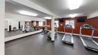 This fitness center is a comprehensive, single-floor facility designed to support a full range of workout routines. The space is organized into distinct functional zones, seamlessly connected to create an open and practical layout. The cardio area is a standout feature, equipped with treadmills, an elliptical trainer, and a stationary bike, all positioned in front of a large floor-to-ceiling mirror that visually expands the room. Bold accent walls in deep red add energy and visual contrast to the otherwise clean white walls and textured ceilings. A mounted flat-screen television provides entertainment during workouts, while windows allow natural light to filter in. The strength training area includes a multi-station cable machine offering a versatile range of resistance exercises, complemented by a well-stocked dumbbell rack spanning a variety of weights. Throughout the facility, durable dark rubber flooring provides safety and comfort underfoot. The space is kept clean and functional with a sanitation station and waste receptacle conveniently located within the facility. Bright overhead flush-mount lighting ensures the entire space is well-illuminated. Multiple doors provide access to additional rooms, including what appear to be restroom or changing facilities, enhancing the overall utility of this fitness center.