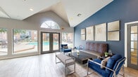 Located at 10750 Westbrae Pkwy in Houston, Texas, this single-floor community clubhouse offers a thoughtfully designed open-plan layout combining a living room, dining area, and kitchen. The space is defined by soaring vaulted ceilings and bold navy blue accent walls that create a striking contrast against the neutral gray tones throughout. Wood-look tile flooring runs seamlessly across the entire space, lending a warm, cohesive feel.

The living area is furnished with contemporary seating, including a sleek gray sofa and royal blue accent chairs with chrome frames, arranged around a dark wood coffee table atop a neutral area rug. A curated gallery wall of framed artwork adds visual interest to the feature wall. French doors with sidelights open to the exterior, filling the space with natural light.

The dining area is bathed in sunlight through expansive floor-to-ceiling windows that frame direct views of the outdoor pool and lush landscaping, creating a bright and inviting atmosphere. High-top tables with metal stools provide casual seating alongside a wall-mounted flat-screen TV.

The kitchen features rich dark wood cabinetry, granite countertops, an undermount sink, and a coffee station, complemented by two framed autumn-themed artworks on the navy accent wall. A breakfast bar with upholstered counter stools connects the kitchen to the main gathering areas.