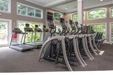 Located at 4000 McGinnis Ferry Rd in Suwanee, Georgia, this impressive fitness center is housed within a single-floor facility defined by its striking architectural character. The centerpiece of the space is a soaring vaulted ceiling supported by bold, exposed wood beam trusses that lend a warm, lodge-inspired aesthetic to the otherwise modern, functional interior. Large windows line the walls and allow abundant natural light to flood the open floor plan, creating an energizing and welcoming workout environment.

The main workout area is expansive, offering generous open floor space designed to accommodate a wide variety of training activities. A ladder agility track is marked directly on the dark commercial-grade carpet, supporting functional fitness and conditioning drills. A heavy-duty multi-station steel rig runs along the center of the space, providing pull-up bars and suspension training anchor points. Along the perimeter, full-length mirrors amplify the sense of space and allow for form monitoring during exercise.

The facility is thoroughly equipped with a comprehensive selection of strength machines, free weights, benches, and a dedicated row of cardio equipment including ellipticals and other aerobic machines. Wall-mounted televisions are positioned throughout for entertainment. The space conveys a professional, well-appointed gym atmosphere suited for a broad range of fitness goals and training styles.