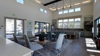 This thoughtfully designed single-floor communal space combines comfort and functionality in a bright, open-concept layout. The room commands attention with its dramatically high ceilings accented by dark exposed beams and a recessed tray ceiling with ambient lighting, lending an air of sophistication to the overall design.

Natural light floods the interior through an expansive array of large windows and clerestory windows positioned along the upper walls, offering views of the surrounding outdoor areas and creating an inviting, airy atmosphere. Glass panel doors open to an outdoor area, further enhancing the connection between interior and exterior.

The space is divided into distinct yet seamlessly connected zones. A casual seating area features contemporary lounge chairs and a low coffee table arranged atop a bold geometric-patterned area rug over wood-look flooring, providing a comfortable spot for relaxed conversation or leisure. Nearby, a dining and collaboration zone offers a rectangular table with upholstered side chairs, ideal for group gatherings or working sessions.

Along one wall, a counter-height bar-style workspace with built-in shelving serves as a dedicated workstation area, accompanied by a stainless steel refrigerator. Pendant lighting above adds a refined finishing touch. The neutral color palette, punctuated by blue-gray tones and organic accents, creates a welcoming and professional ambiance throughout.