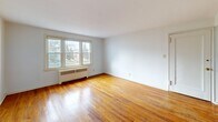 Located at 251 Hillcrest Manor Ct in Utica, NY, this 4-bedroom, 1-bathroom unit offers a practical and well-organized single-floor layout. Warm, polished hardwood floors run consistently throughout the bedrooms and hallway, lending a sense of continuity and classic character to the space. Each bedroom is bright and simply appointed, with white walls, natural light streaming through double-hung windows, and radiator heating units ensuring year-round comfort. The spacious primary bedroom stands out with multiple windows offering outdoor views of the surrounding neighborhood. A connecting hallway links the rooms seamlessly, as seen in the dollhouse view, with clean white panel doors and brass hardware adding a subtle traditional touch. The kitchen features white cabinetry, a gas range, a single-basin stainless steel sink, wood-toned countertops, and a window overlooking the exterior — functional and straightforward. The bathroom is a notable retro-styled space, finished with pink ceramic wall tiles accented by a bold black border trim, a full soaking tub, and a window providing natural ventilation. The overall ambiance of the unit is clean, bright, and functional, offering a comfortable and classic living environment.