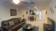 This two-floor residence offers a thoughtfully designed layout that balances open communal spaces with practical functionality. The main living area is anchored by plush carpeting, a ceiling fan, and recessed lighting that together create a warm, well-lit atmosphere. A comfortable seating arrangement centers around a round coffee table, providing an inviting space for relaxation and entertaining.

Adjacent to the living room, a dedicated dining area accommodates a classic table-and-chair set, seamlessly bridging the gap between the lounge and kitchen spaces. The kitchen features a pass-through counter that opens directly to the dining area, enhancing the open-concept feel and allowing for easy interaction between spaces. Wood-toned cabinetry lines the kitchen walls, complemented by recessed lighting overhead and modern black appliances.

Natural light filters into the space through a set of vertical blinds near the entryway, brightening the neutral-toned walls and light carpet throughout. The overall ambiance is clean, cohesive, and move-in ready, with a color palette of warm beiges and browns that lends a grounded, comfortable character to the interior. The two-floor layout provides additional living or sleeping space beyond the main level, adding versatility to the overall floor plan.