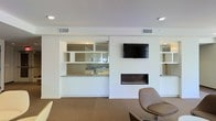 This single-floor space showcases a clean, contemporary design defined by an all-white interior palette and thoughtful architectural details. The main living area serves as the heart of the space, anchored by a sleek, linear built-in fireplace set into a crisp white accent wall. A wall-mounted flat-screen television sits just above the fireplace, creating a streamlined and functional focal point for the room. Flanking the feature wall are open built-in shelving units, offering both display and storage opportunities while maintaining the space's minimalist aesthetic. Adjacent to the living area, a pass-through kitchen window opens seamlessly into the main room, promoting an open and connected feel. The kitchen features light granite or stone countertops and is equipped with a sink, with mirrored backsplash elements that reflect light and add depth to the space. Recessed lighting throughout the ceiling provides even, ambient illumination, complementing the neutral carpeted flooring that adds warmth underfoot. The overall ambiance is polished and understated, with a cohesive color scheme of whites, grays, and warm neutrals that lends the space a sophisticated yet inviting character.