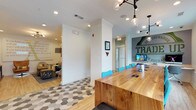 This single-floor space showcases a contemporary and well-curated interior design that blends functionality with style. The open-plan layout creates a seamless flow between a collaborative workspace and a lounge area, making it ideal for both focused work and casual interaction. A standout feature is the long, solid natural wood communal table paired with bright teal chairs, anchored beneath a striking industrial-style pendant light fixture with exposed Edison bulbs — a combination that balances warmth and modernity. The workspace is equipped with integrated power and connectivity ports along the table, complemented by a mounted display screen, making it well-suited for meetings and collaborative sessions. The flooring is a distinctive geometric hex-tile pattern in shades of grey and white, adding a bold visual element to the space. Adjacent to the main workspace, a lounge area provides comfortable seating amid softer lighting and décor. The walls feature motivational typography and branded graphics, lending the space an energetic, community-driven character. Recessed lighting throughout ensures the interior is well-lit, while white crown molding and structural columns add a refined architectural touch. The overall ambiance is modern, inviting, and purpose-driven.
